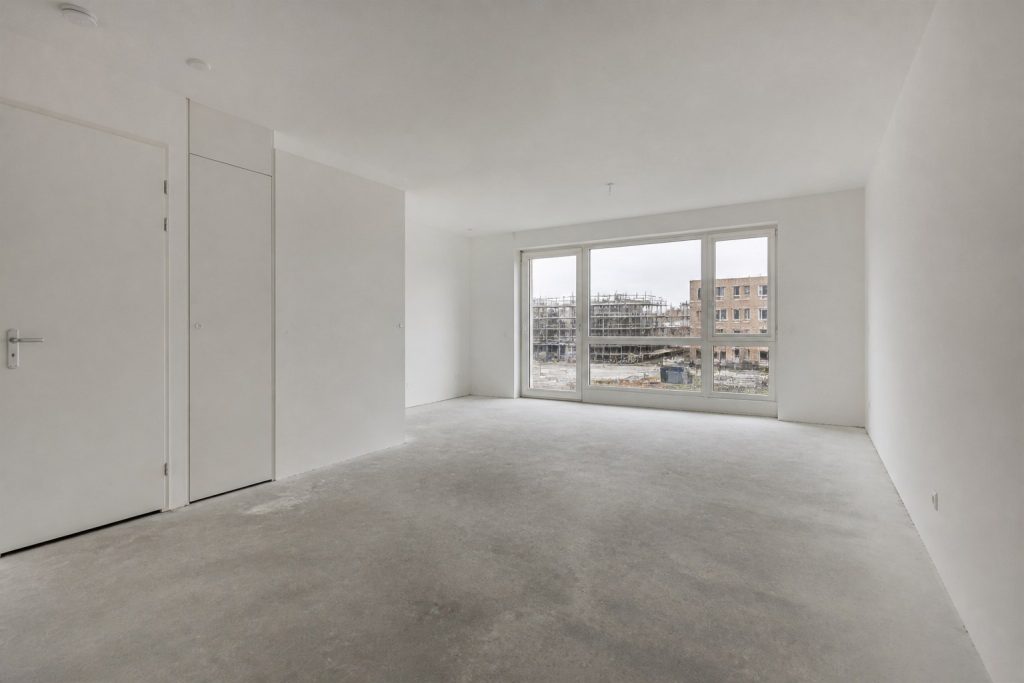Lege woonkamer van een nieuwbouwappartement met kale dekvloer, witte wanden en uitzicht op een bouwplaats