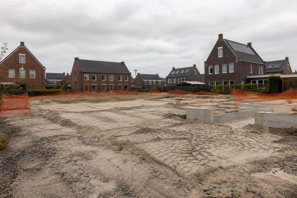 Leeg bouwrijp terrein met zandsporen, oranje bouwhekken en betonnen funderingsblokken naast afgebouwde woningen